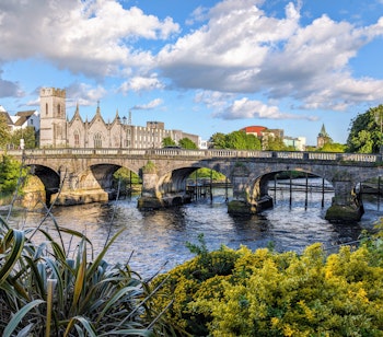 Den historiske Salmon Weir bro i Galway med katedralen og byens bygninger i baggrunden ved floden Corrib under blå himmel