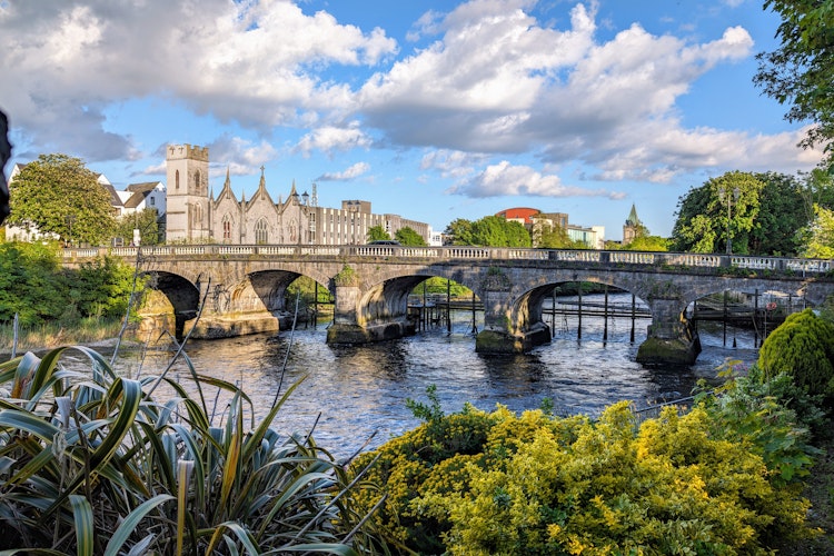 Den historiske Salmon Weir bro i Galway med katedralen og byens bygninger i baggrunden ved floden Corrib under blå himmel