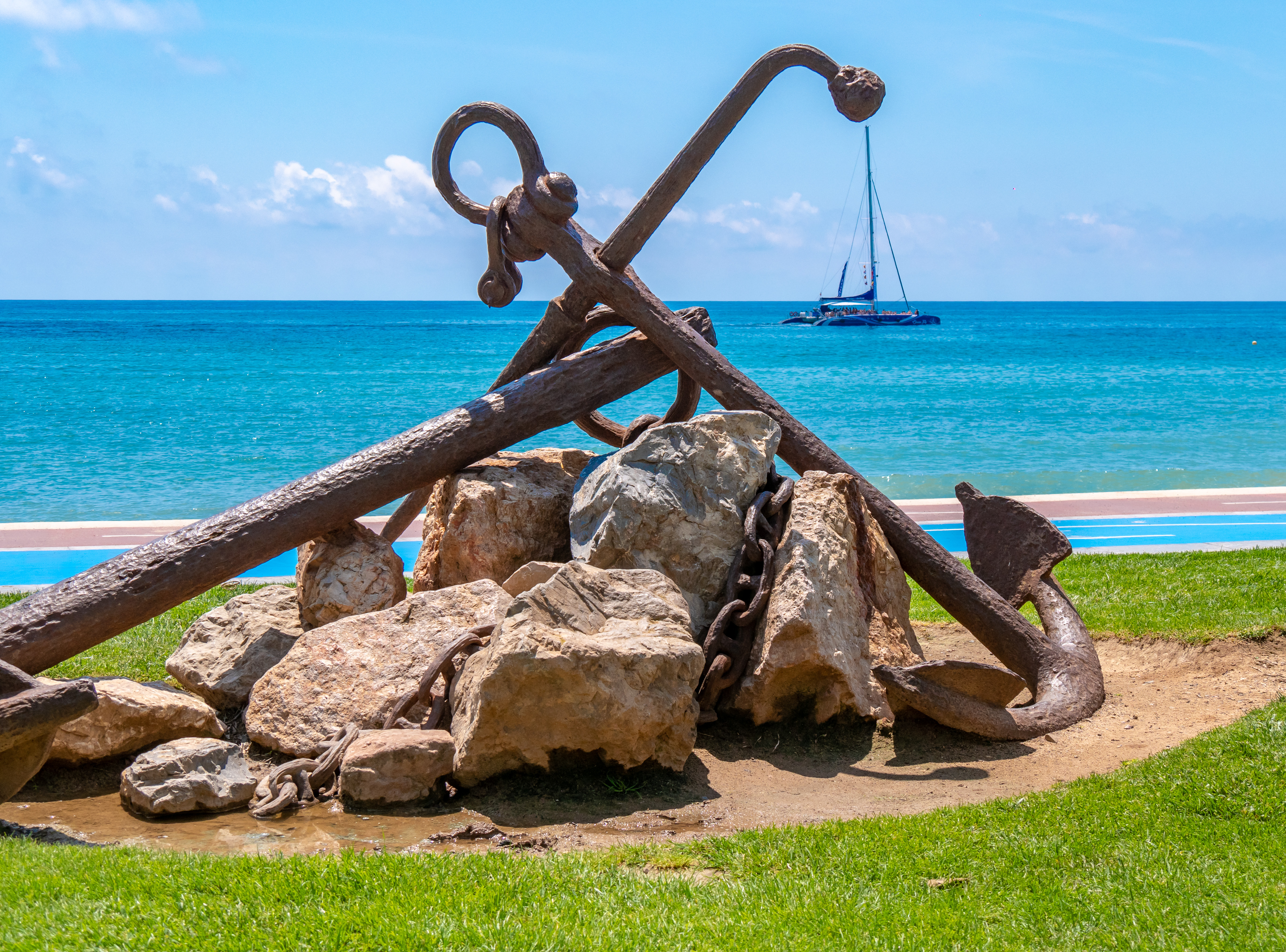 Historisk skibsanker monument ved Middelhavskyst med sejlbåd i baggrunden og turkisblåt vand