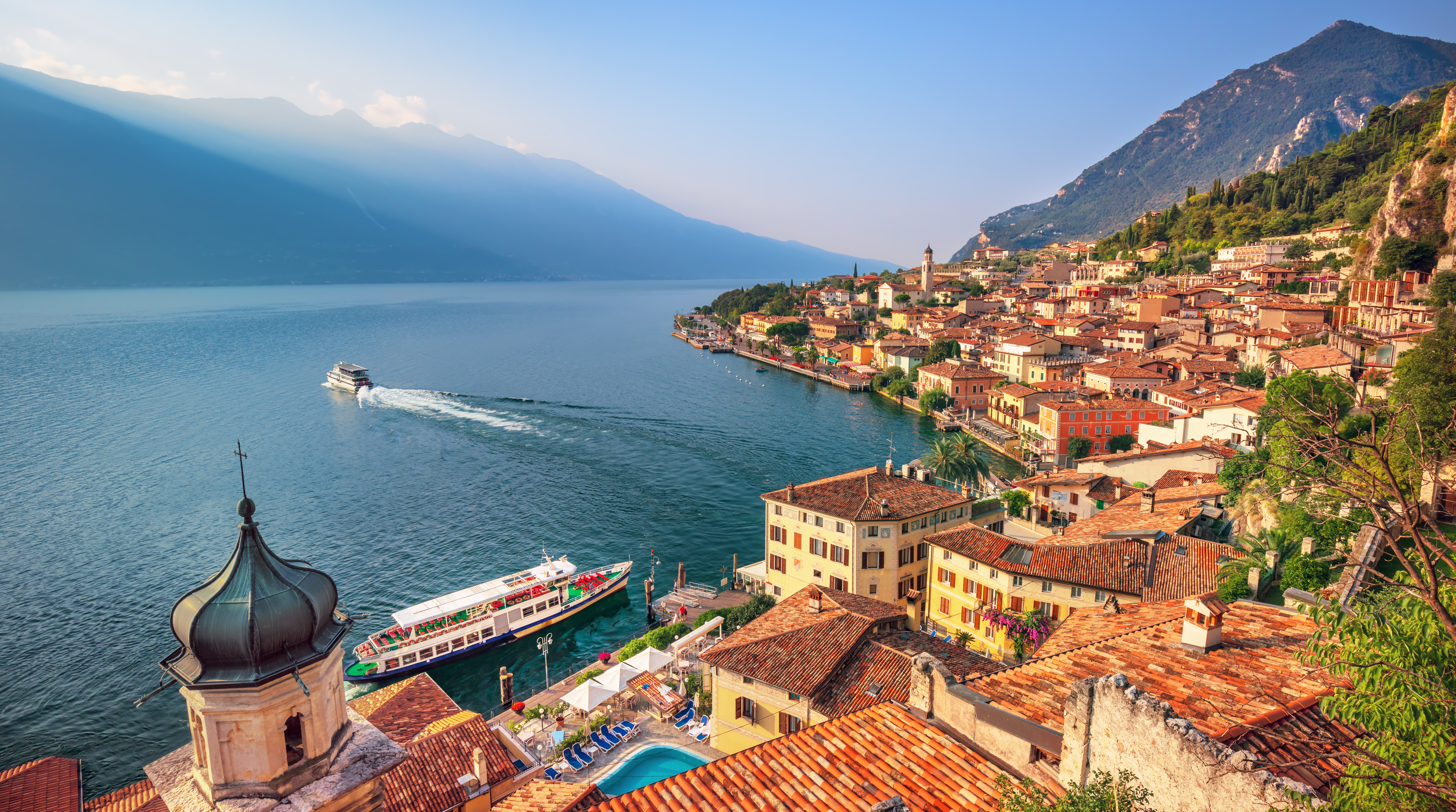 Smuk udsigt over Gardasøen med historisk italiensk by, farverige bygninger, kirketårn og færger på det azurblå vand omgivet af bjerge