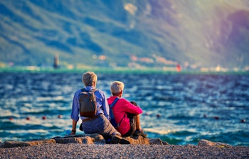 To rejsende sidder ved Gardasøens bred i efteråret og nyder den storslåede udsigt over det azurblå vand omgivet af majestætiske bjerge i Trentino Alto Adige regionen