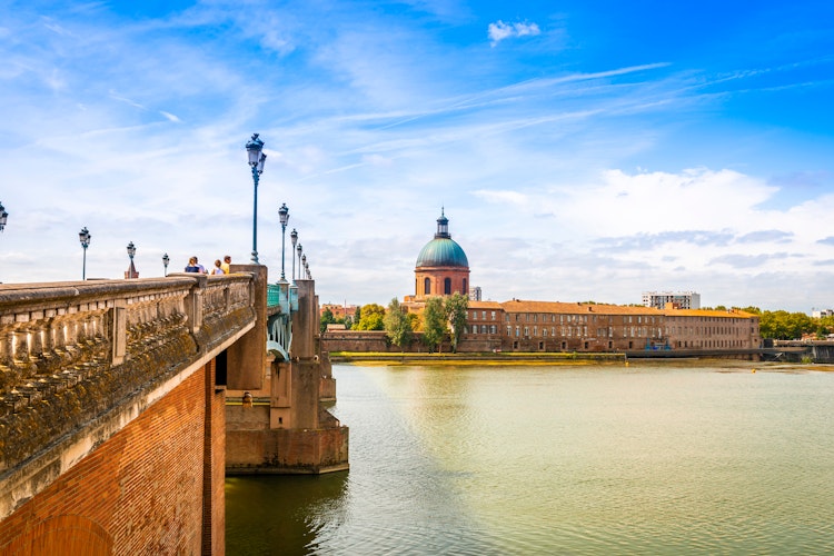 Garonne-floden med historiske broer og Toulouses smukke skyline ved solnedgang, hvor den lyserøde bys bygninger spejler sig i vandet under den blå aftenhimmel