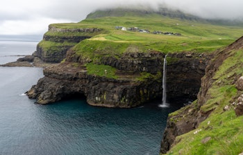 Betagende luftfoto af Gasadalur vandfald der falder direkte i havet med den idylliske landsby omgivet af grønne fjelde på Færøerne