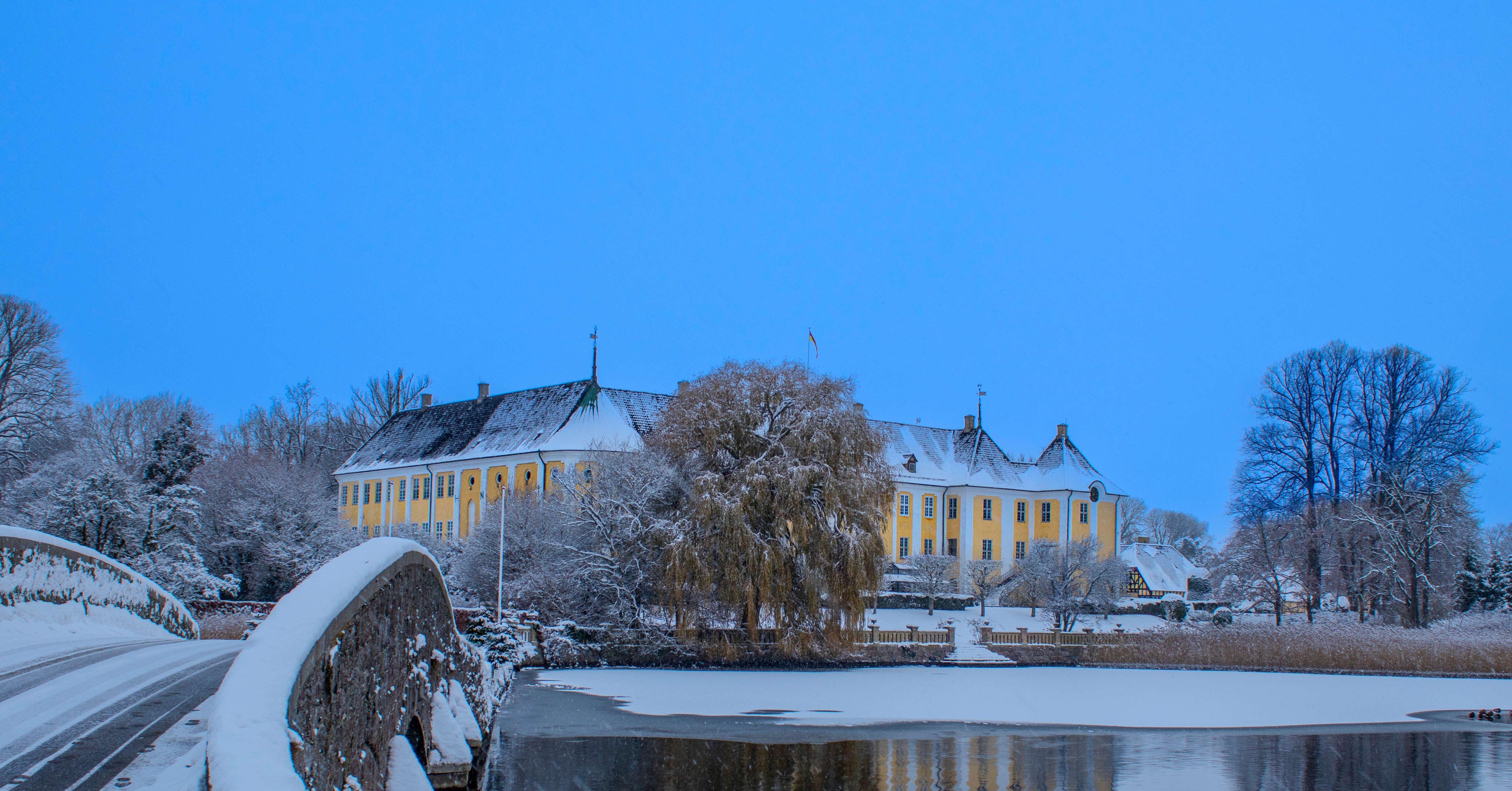 Gavnø Slot oplyst i vinterlandskab med sne, frossen sø og stenbro i forgrunden ved blå vinterhimmel