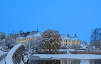 Gavnø Slot oplyst i vinterlandskab med sne, frossen sø og stenbro i forgrunden ved blå vinterhimmel