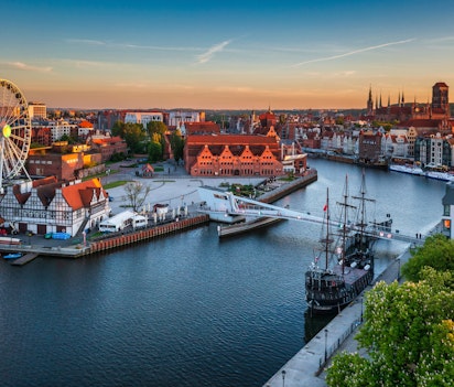 Luftfoto af Gdansks gamle bydel ved solnedgang med pariserhjul, historiske pakhuse, farverige bygninger og skibe langs Motlawa-floden i Polen under en orange himmel