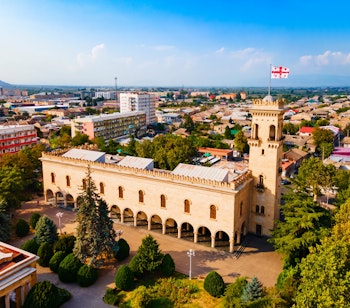 Historisk georgisk fæstning med tårn og buegang omgivet af moderne bybygninger under blå himmel