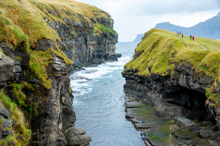 Dramatisk havkløft ved landsbyen Gjógv på Færøerne med turkisblåt atlanterhavsvand der bryder mod de stejle grønklædte klipper og turister der nyder udsigten