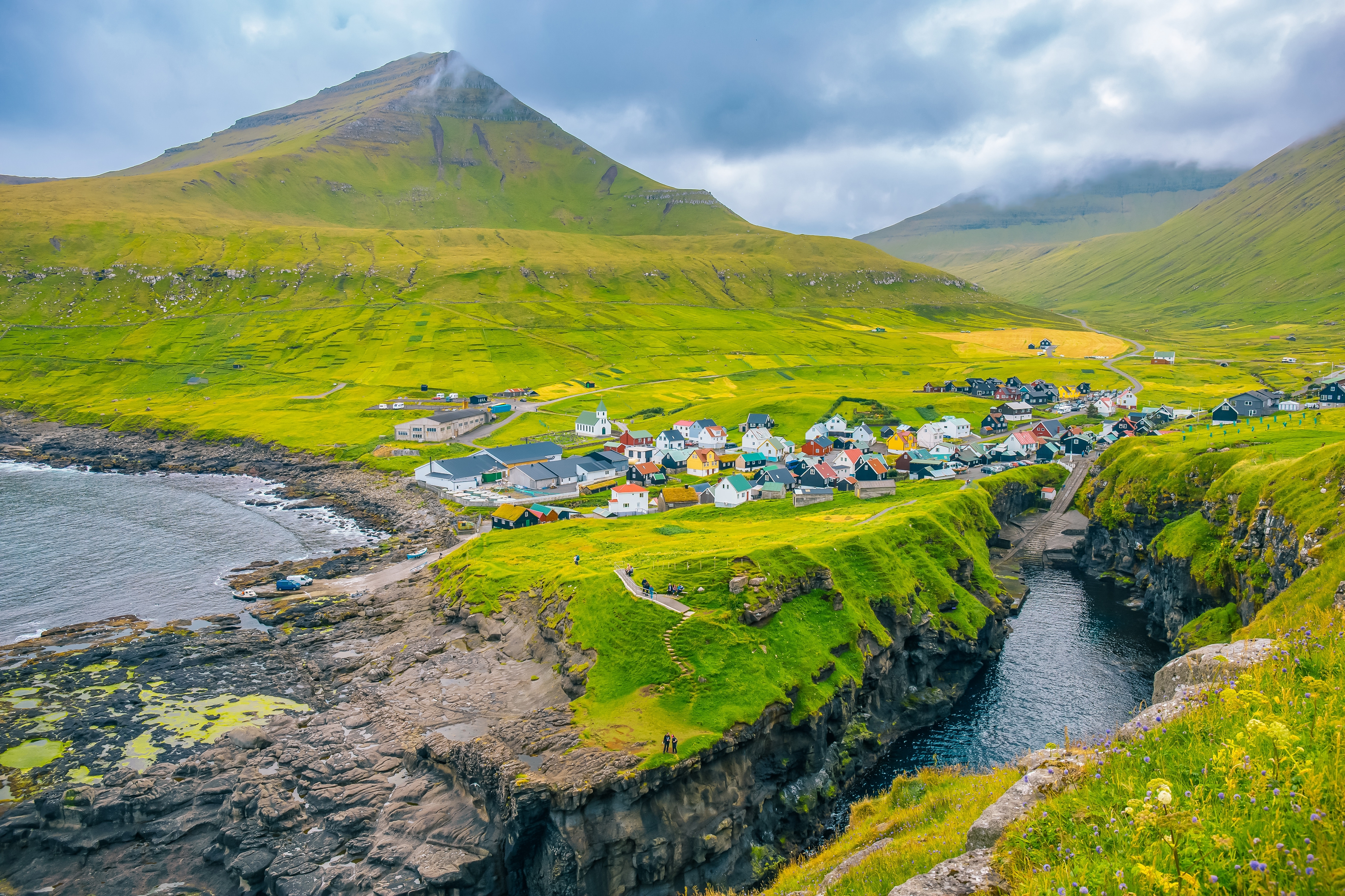 Luftfoto af den farverige landsby Gjógv på Eysturoy på Færøerne, omgivet af grønne klippekyster, en naturlig havnegorge og et majestætisk bjerg i baggrunden