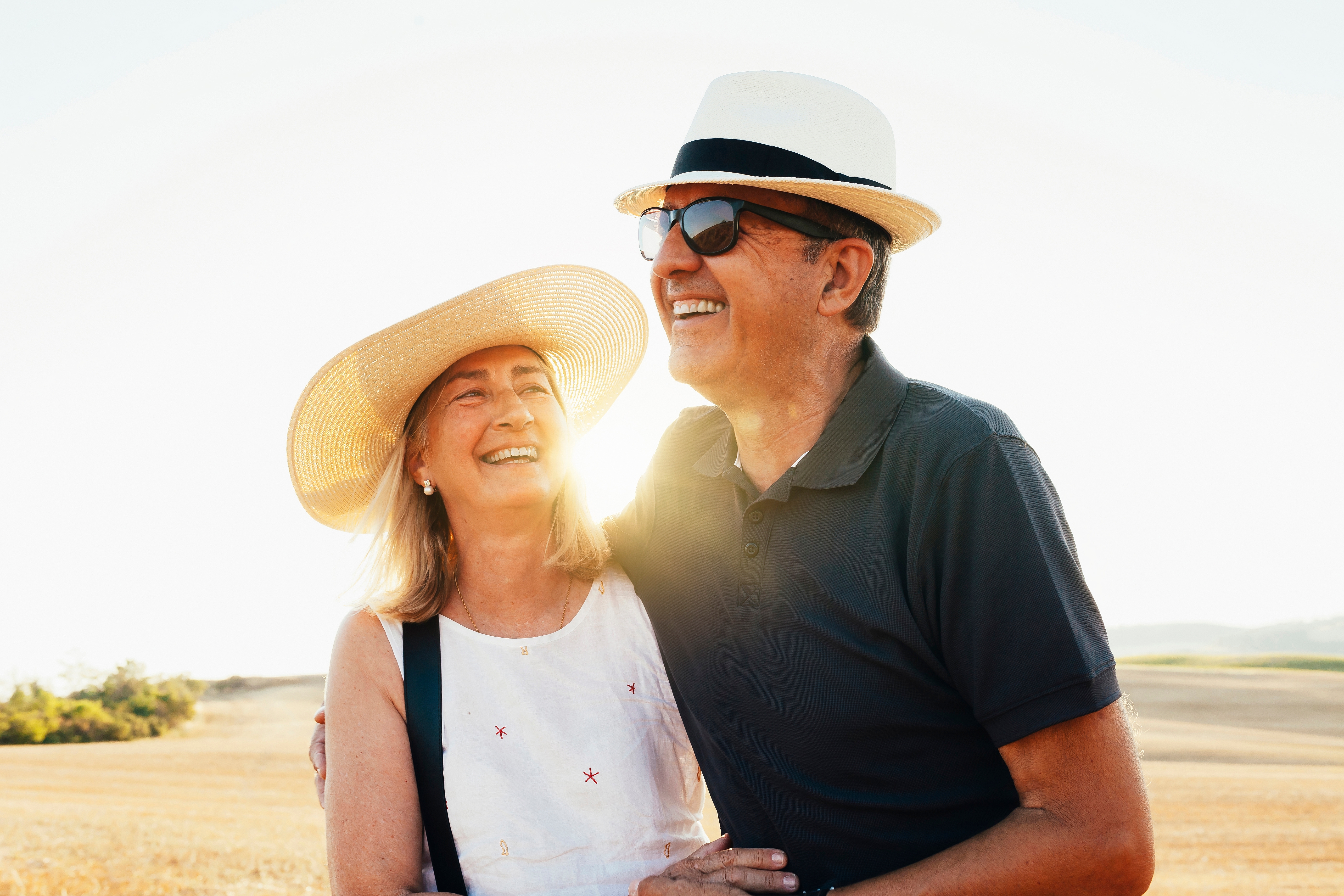 Glad seniorpar i hatte og solbriller omfavner hinanden i gyldent toscansk mark ved solnedgang i Italien