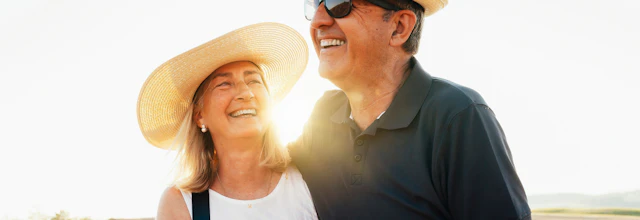 Glad seniorpar i hatte og solbriller omfavner hinanden i gyldent toscansk mark ved solnedgang i Italien