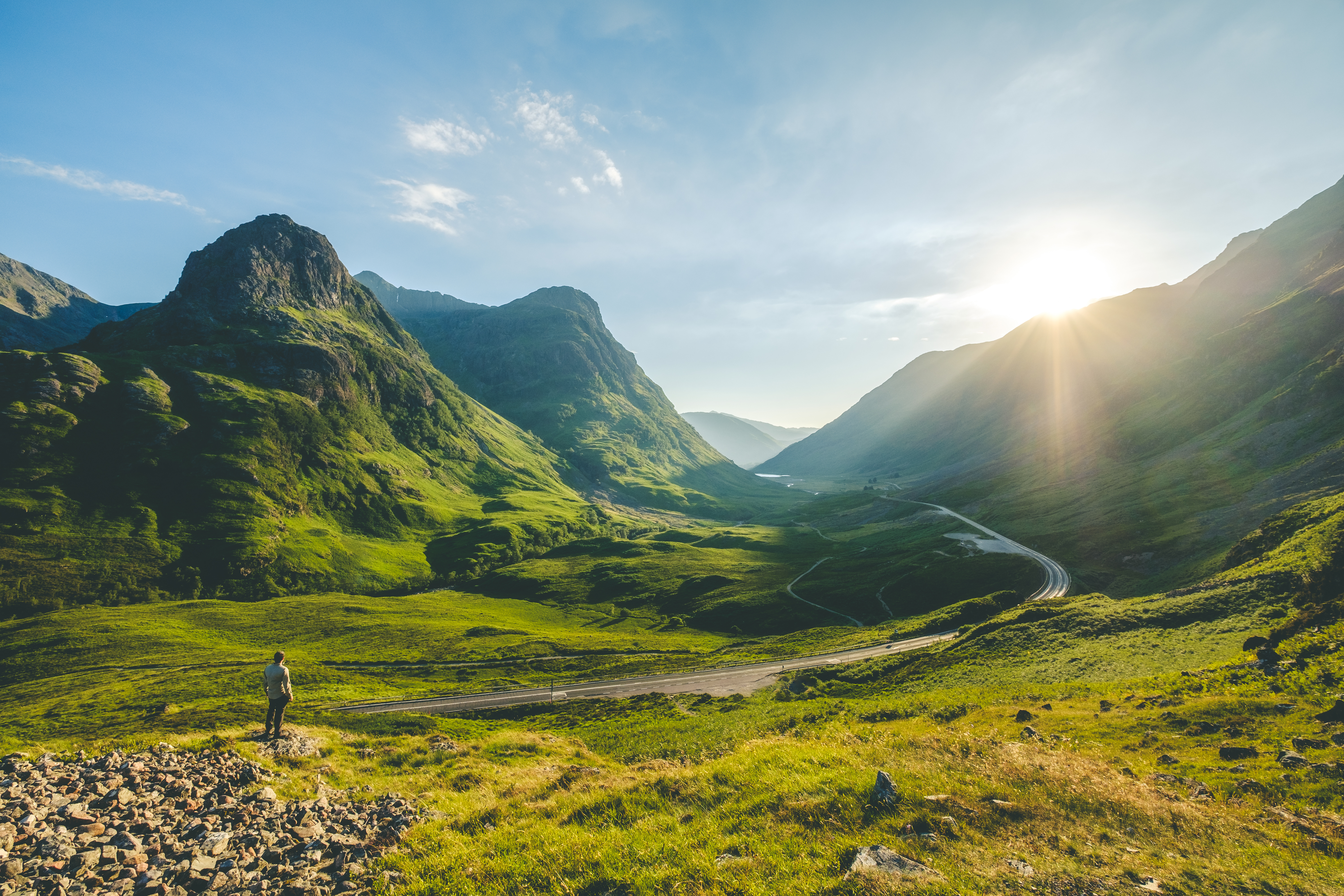 Vandrер står og nyder udsigten over den dramatiske Glen Coe dal i Skotland med snoet vej og gyldent sollys bag bjergene