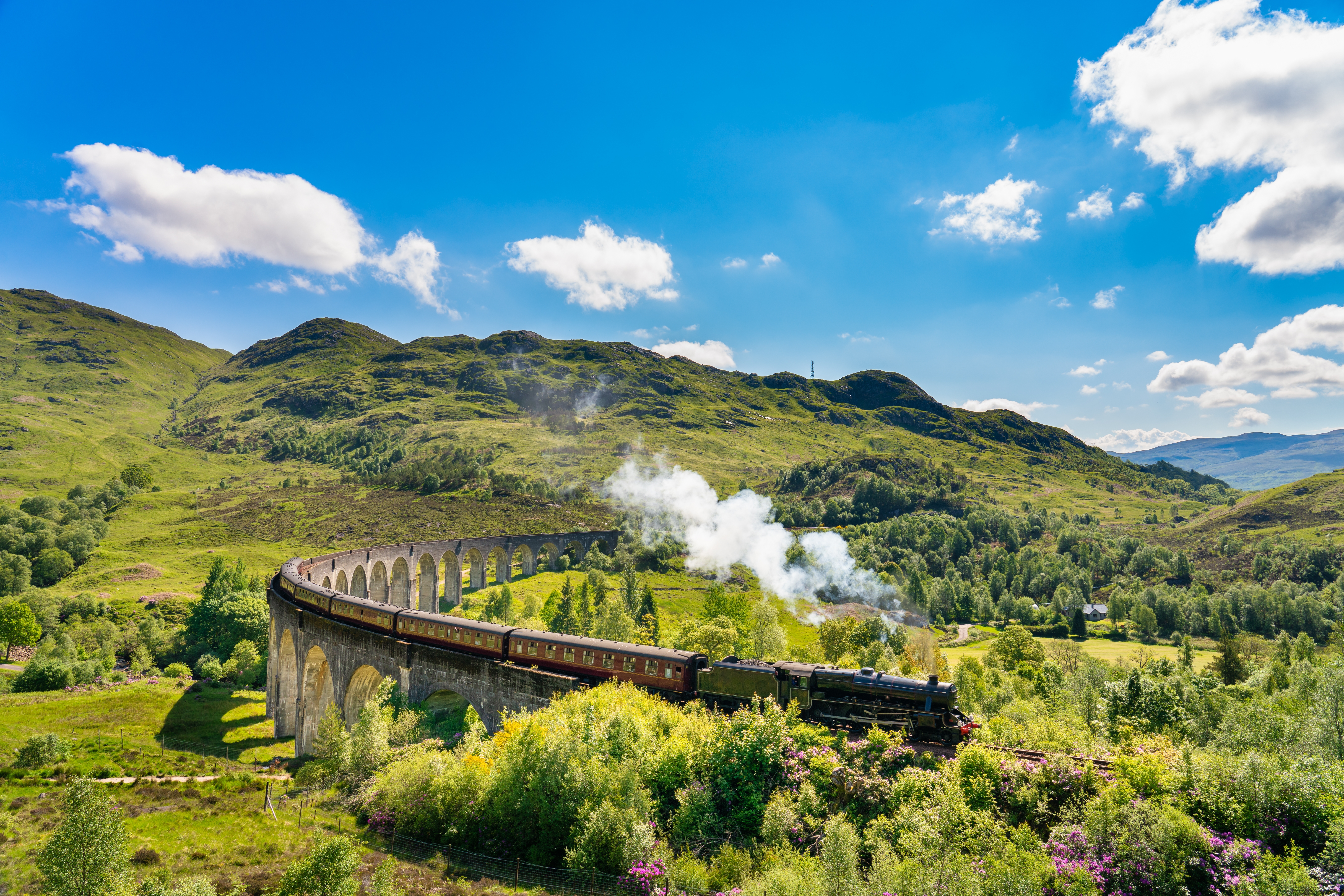 Damptog krydser det berømte Glenfinnan Viadukt i Skotlands Højland med grønne bjerge og blå himmel