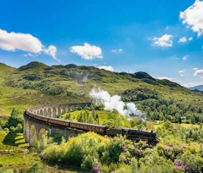 Damptog krydser det berømte Glenfinnan Viadukt i Skotlands Højland med grønne bjerge og blå himmel