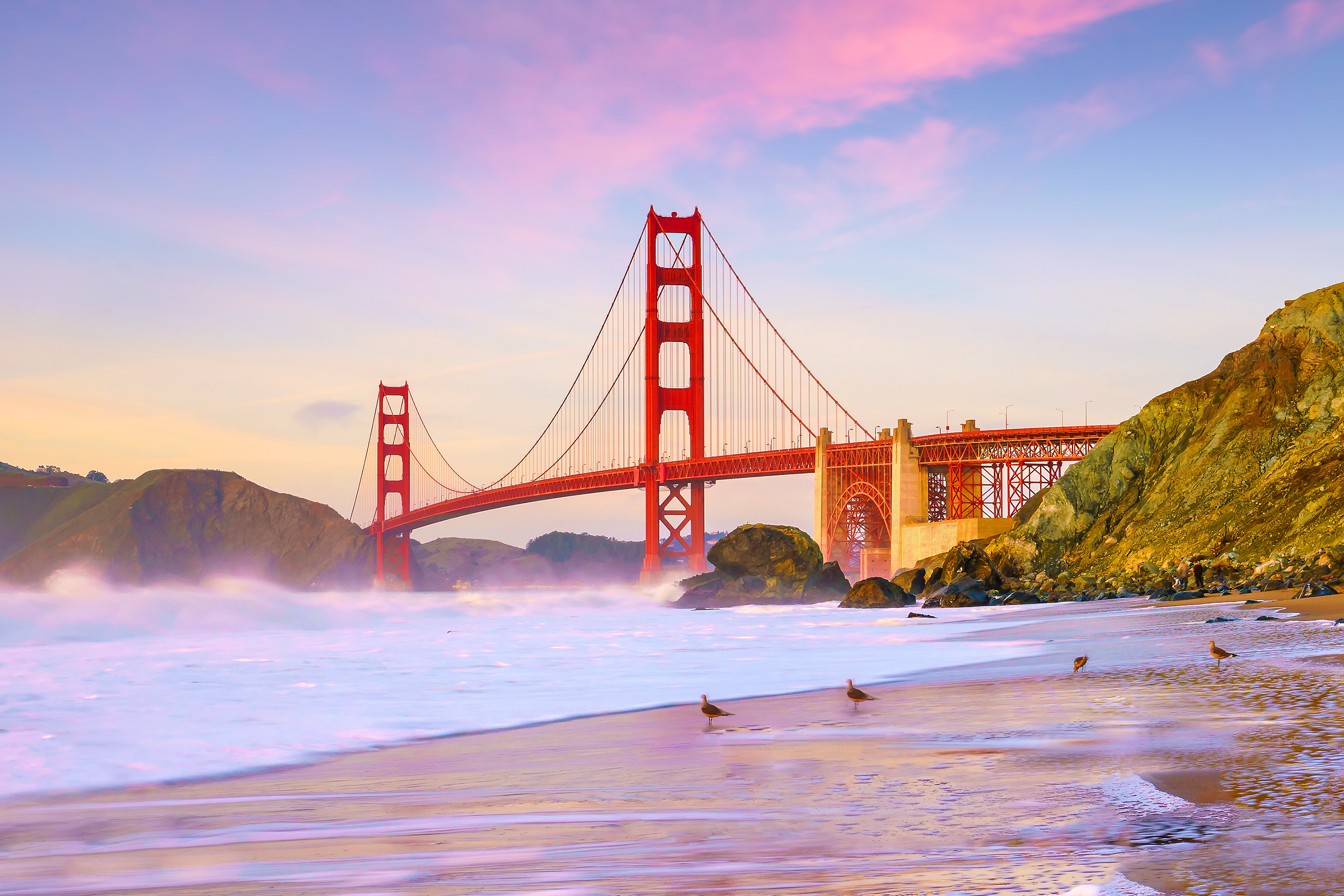 Golden Gate Bridge ved solnedgang set fra stranden i San Francisco, Californien, med lyserød og lilla himmel over Stillehavet