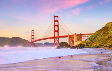 Golden Gate Bridge ved solnedgang set fra stranden i San Francisco, Californien, med lyserød og lilla himmel over Stillehavet