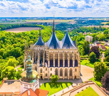 Luftfoto af majestætisk gotisk katedral med tre karakteristiske tårne omgivet af grøn natur i Tjekkiet