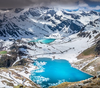 Betagende bjergvej der snor sig omkring en turkisblå alpesø i Gran Paradiso Nationalpark med sneklædte bjergtinder i baggrunden