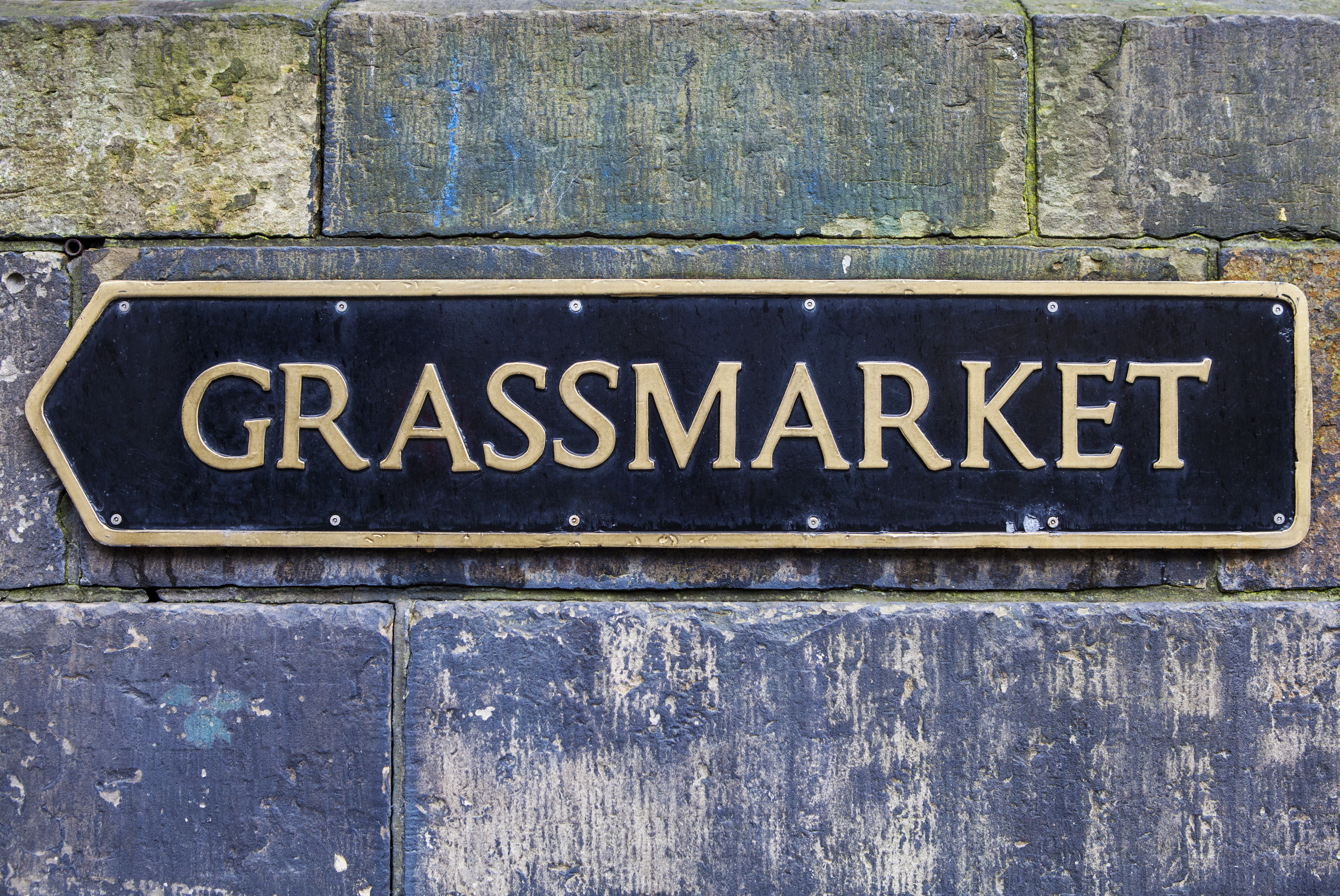 Sort og guldfarvet gadeskilt for Grassmarket monteret på historisk stenmur i Edinburgh, Skotland