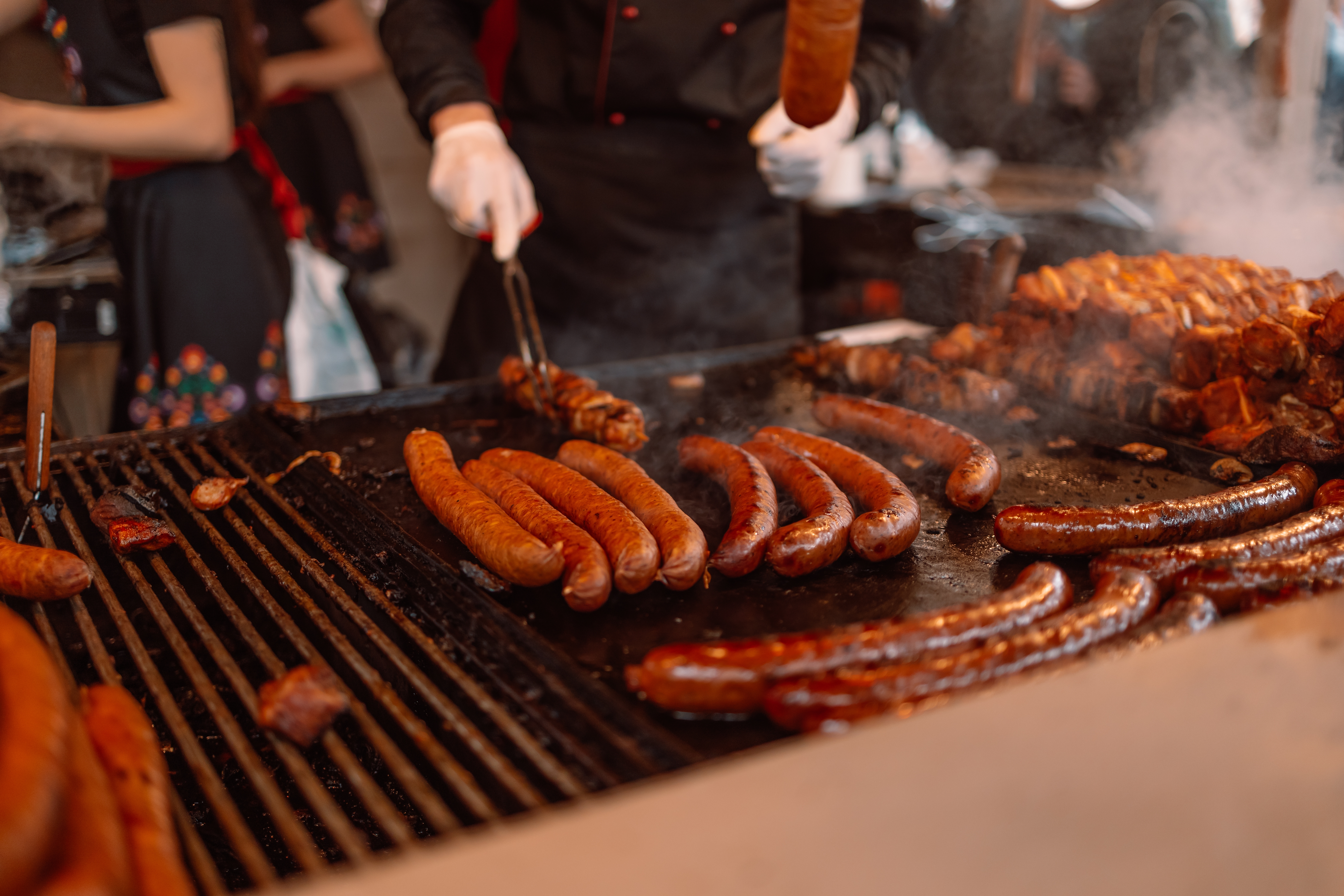 Dampende grillede pølser på en stor udendørs grill ved et street food marked i Krakow, Polen, hvor en kok i sort uniform vender pølserne med en tang