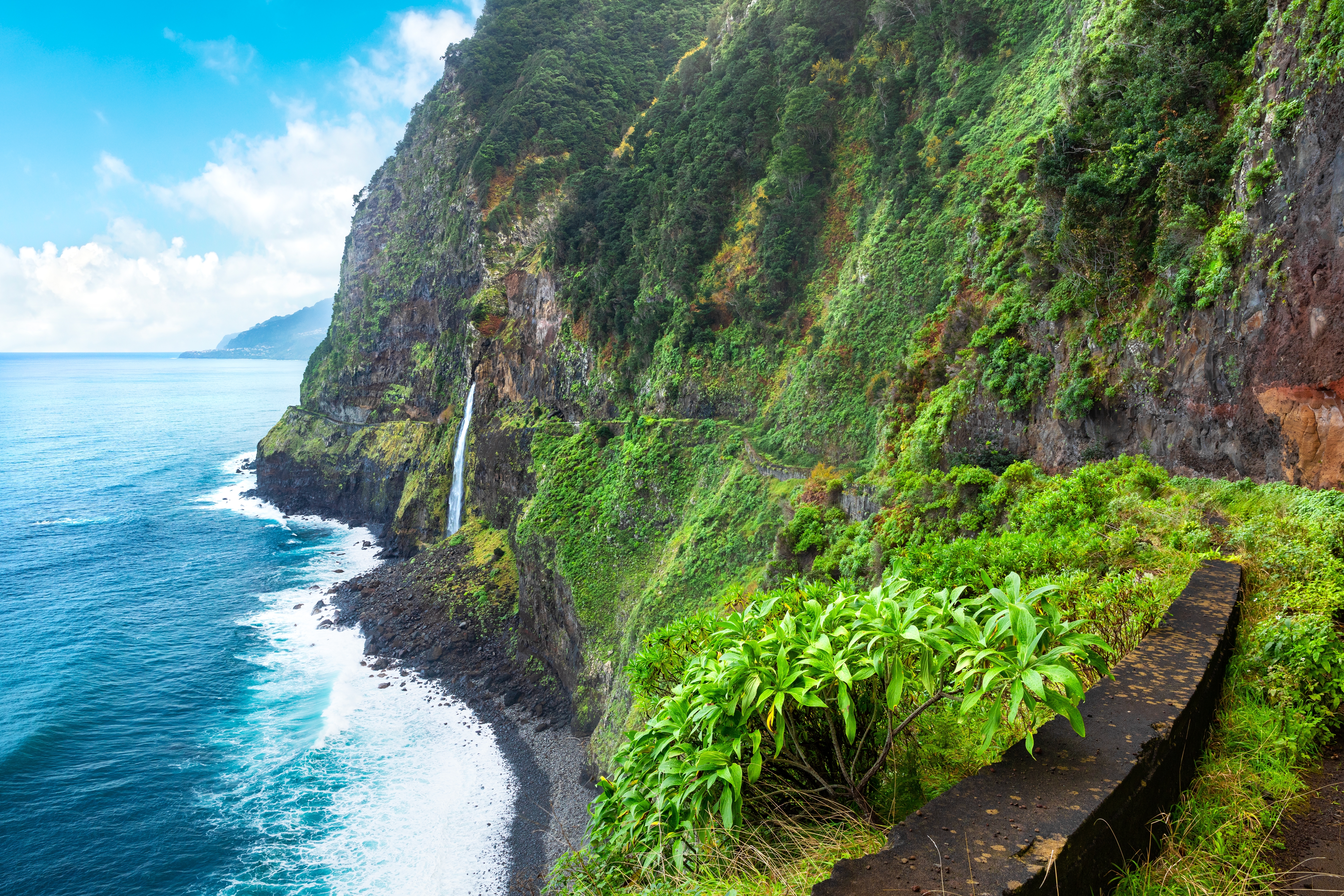 Dramatiske grønne kystskrænter med frodig vegetation og vandfald der styrter ned i turkisblåt hav