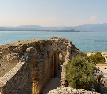 Historiske ruiner fra Grotte di Catullo i Sirmione med udsigt over Gardasøens azurblå vand og bjerglandskab. Resterne af den romerske villa vidner om områdets rige historie