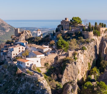 Betagende luftfoto af Guadalest bjergby med historisk slot på klippetop og turkisblå reservoirsø i Costa Blanca, Spanien