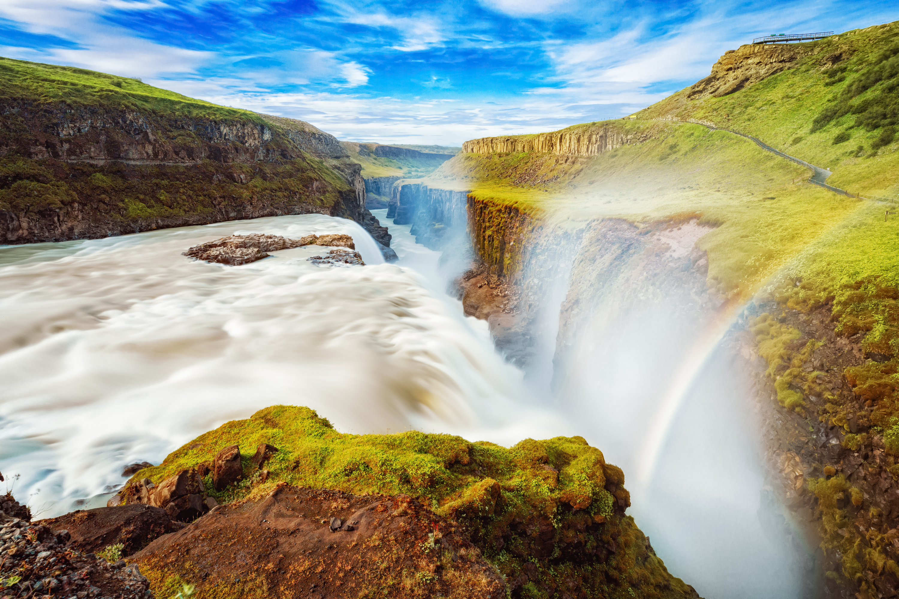 Gullfoss vandfald på Island med regnbue i vandsprøjt og grønne mosbedækkede klipper under blå himmel