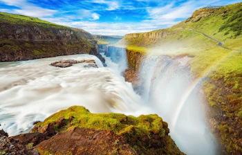 Gullfoss vandfald på Island med regnbue i vandsprøjt og grønne mosbedækkede klipper under blå himmel