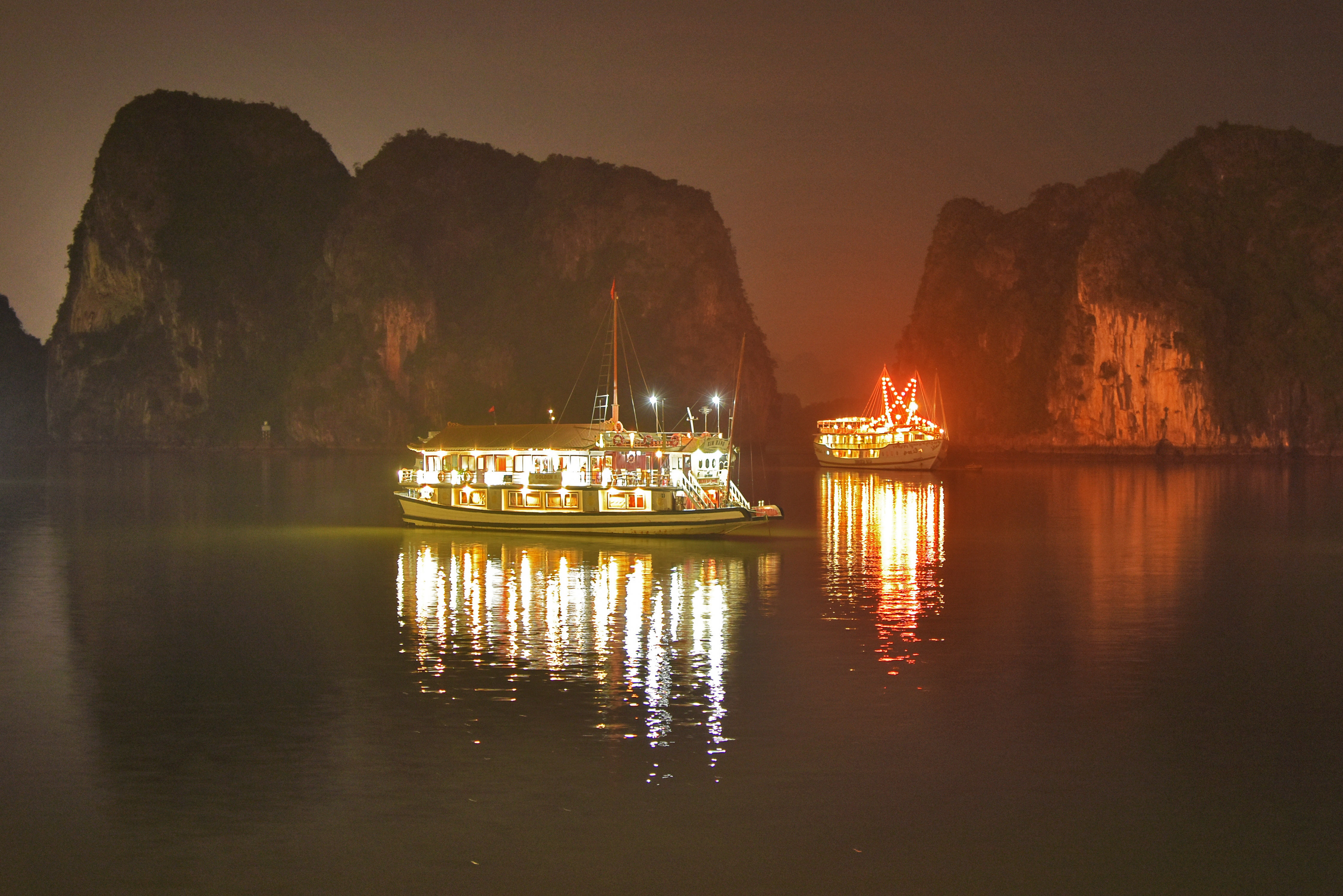 Oplyste cruisebåde i Ha Long Bay Vietnam ved solnedgang med kalkstensformationer og refleksioner i vandet