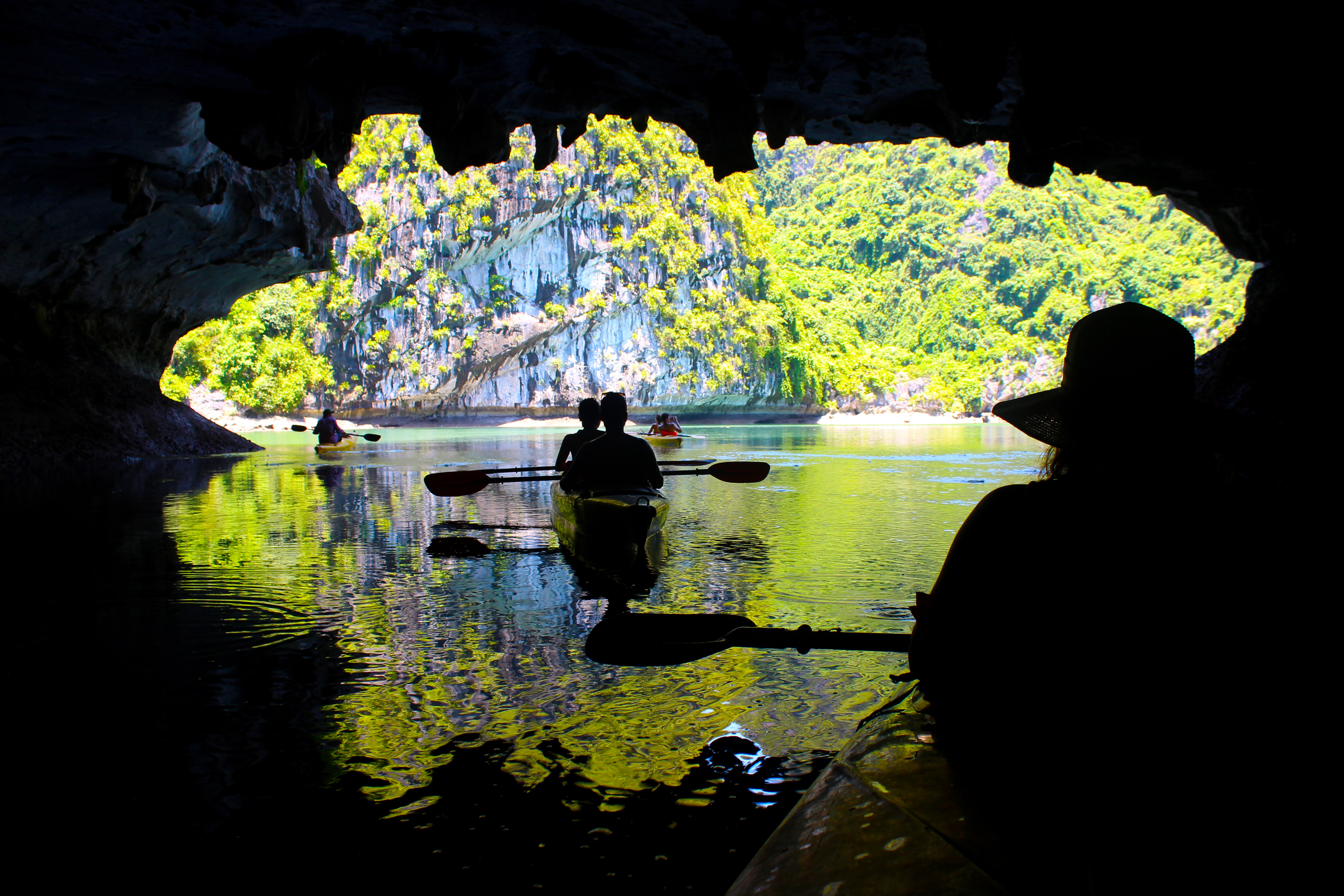 Silhuetter af et par der padler kajak gennem en kalkstensgrotte i Halong Bugten, Vietnam, med en dramatisk kontrast mellem den mørke hule og det turkisblå vand, der skinner ind gennem grotteåbningen