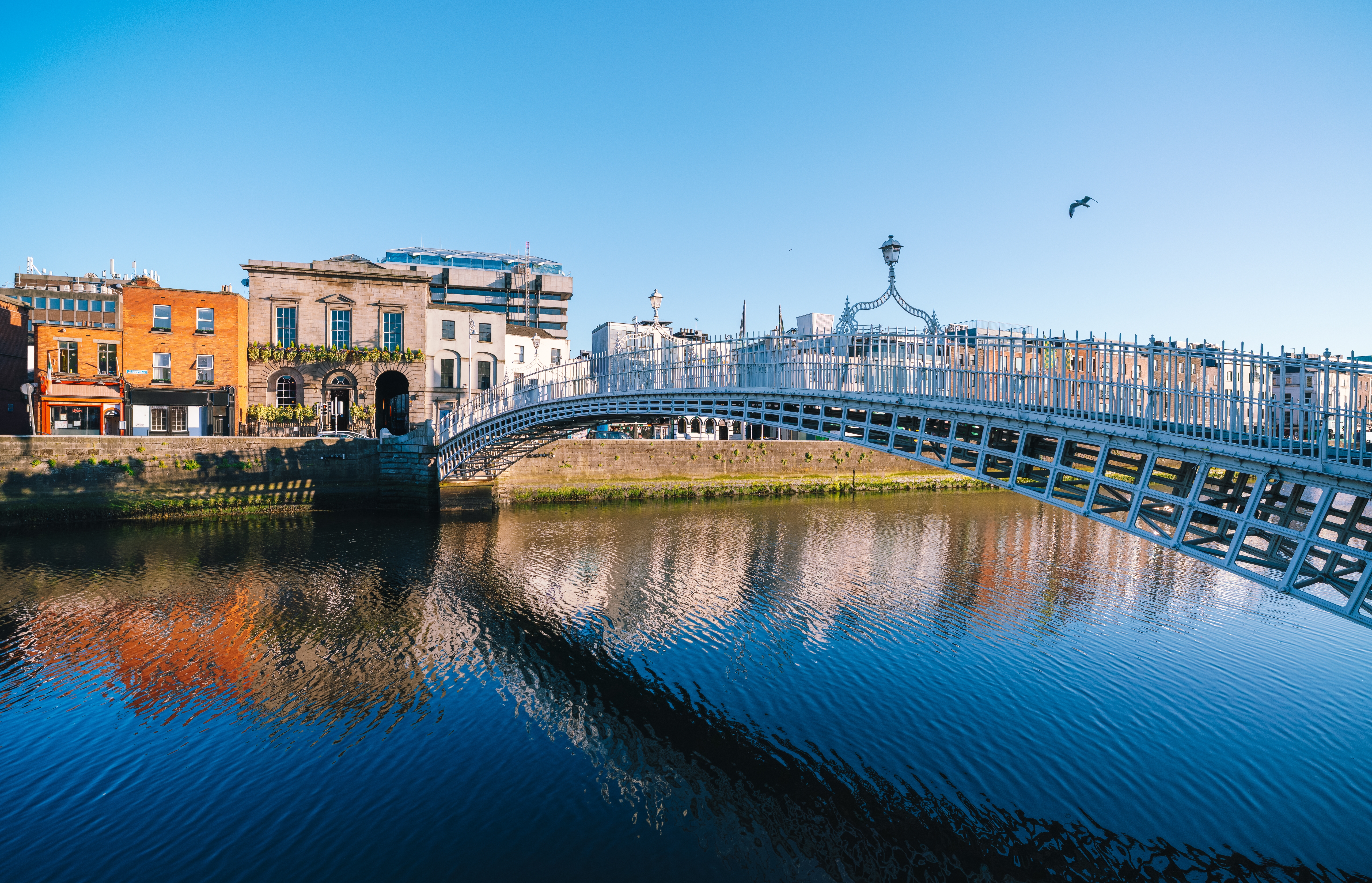 Ha'penny Bridge over floden Liffey i Dublin, Irland, med historiske bygninger der spejler sig i det rolige vand under en klar blå himmel
