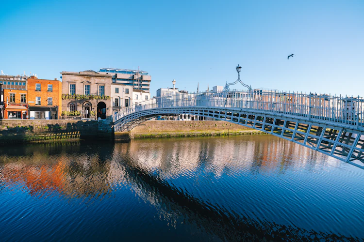 Ha'penny Bridge over floden Liffey i Dublin, Irland, med historiske bygninger der spejler sig i det rolige vand under en klar blå himmel