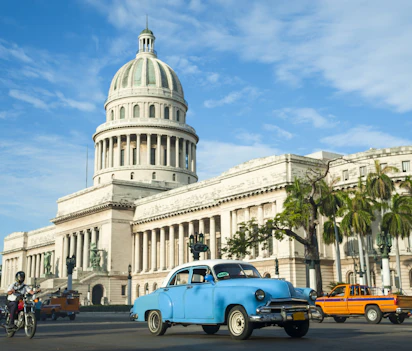 Klassisk blå veteranbil foran El Capitolio bygningen i Havana, Cuba med palmer og blå himmel