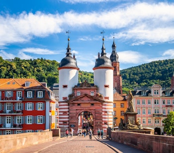 Idyllisk udsigt over den berømte Gamle Bro i Heidelberg med dens karakteristiske tvillingetårne, farverige bygninger i den gamle bydel og de grønne bjerge under en blå himmel