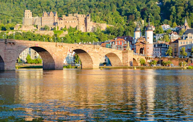 Den historiske Karl Theodor Bro i Heidelberg, Tyskland, med middelalderlige slotsruiner på bakken over Neckar-floden i gyldent aftenlys