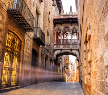 Stemningsfuld neogotisk bro mellem historiske bygninger i Barri Gotic-kvarteret i Barcelona oplyst af gule lamper