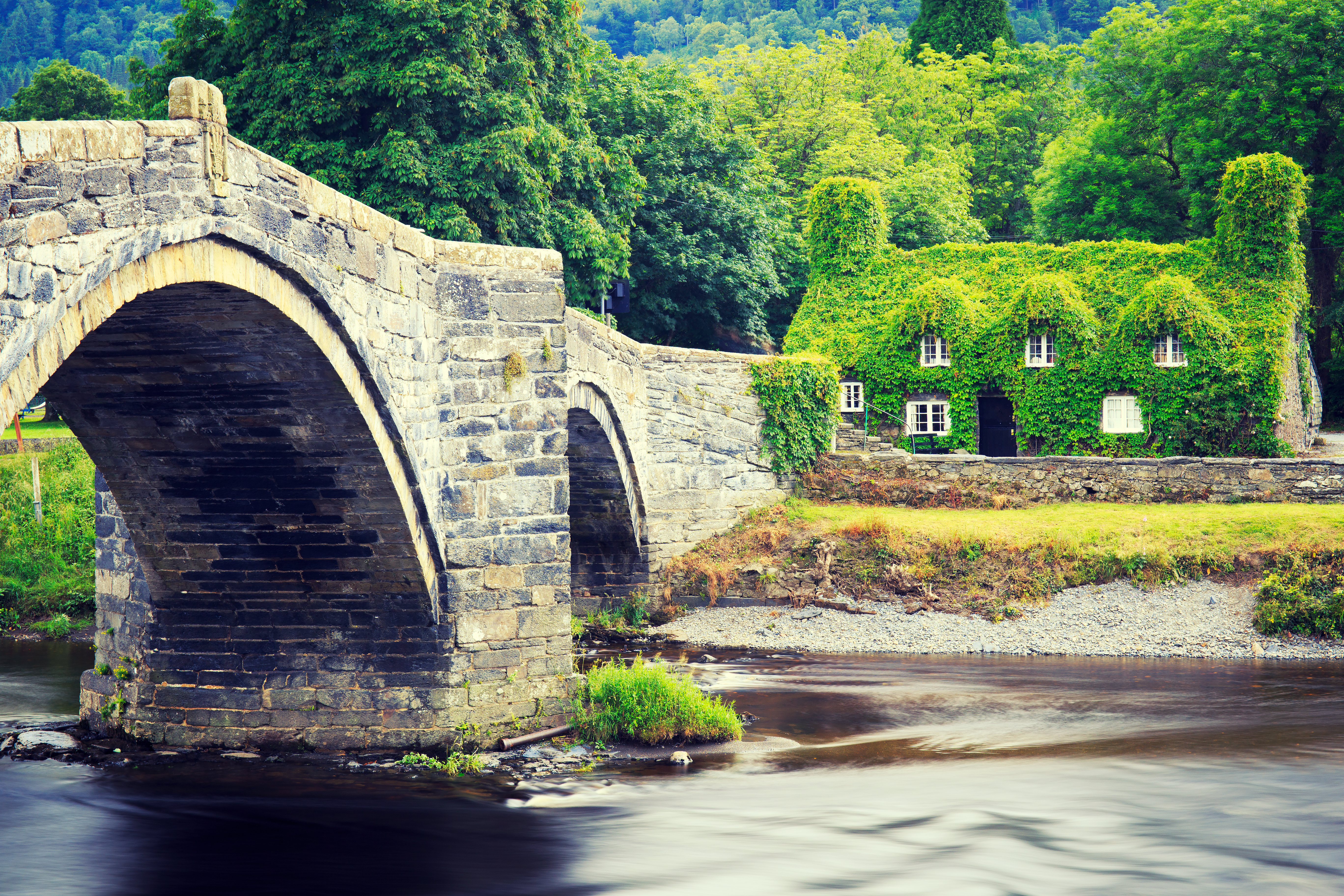 Charmerende historisk stenbro over Conwy-floden med traditionelt vinbeklædt hus i Llanrwst, Nordwales. Idyllisk walisisk landskab med grønne bakker