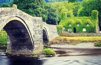 Charmerende historisk stenbro over Conwy-floden med traditionelt vinbeklædt hus i Llanrwst, Nordwales. Idyllisk walisisk landskab med grønne bakker