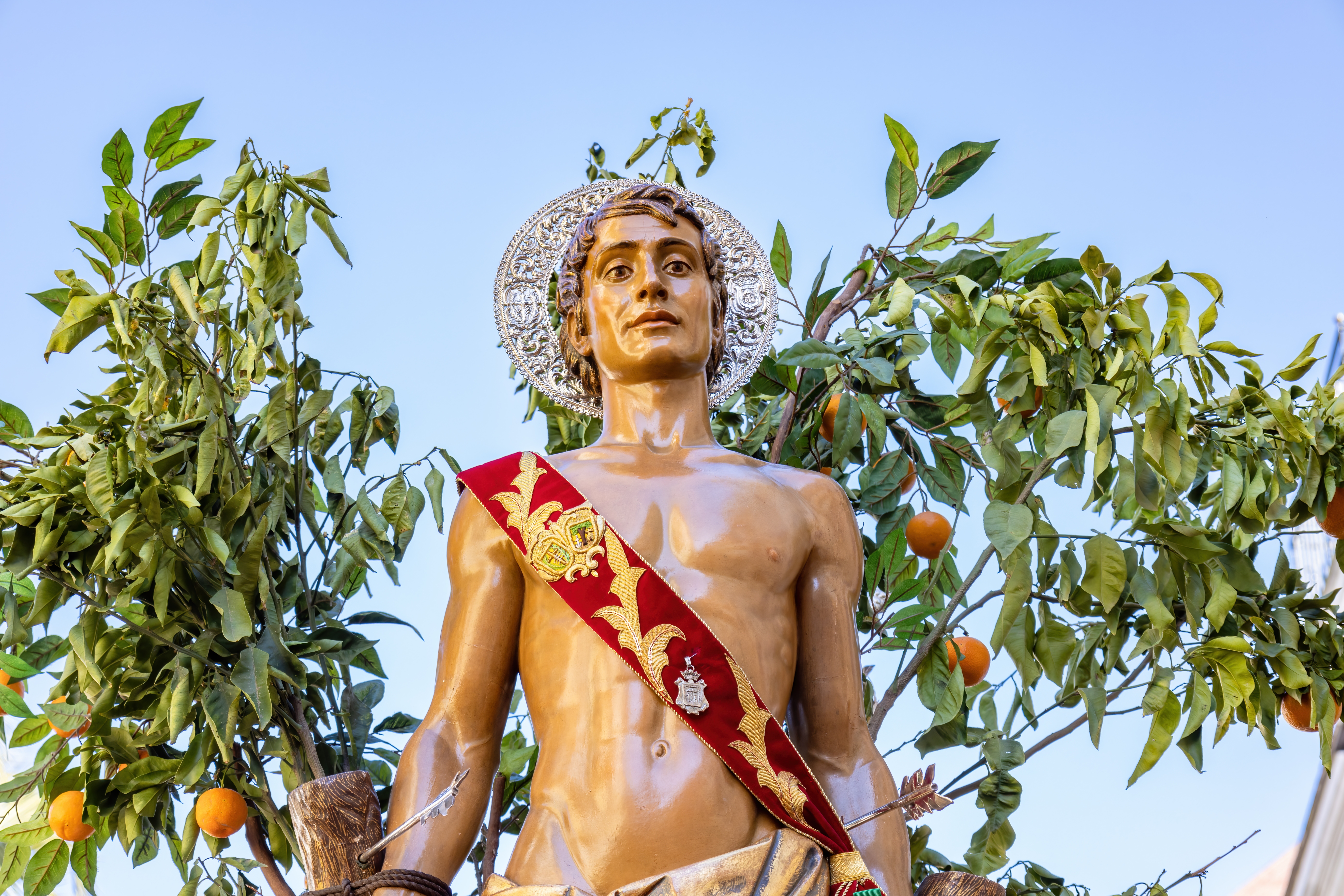 Detaljeret statue af skytshelgenen San Sebastian båret på en blomsterdekoreret platform under en traditionel religiøs procession i Huelvas historiske gader