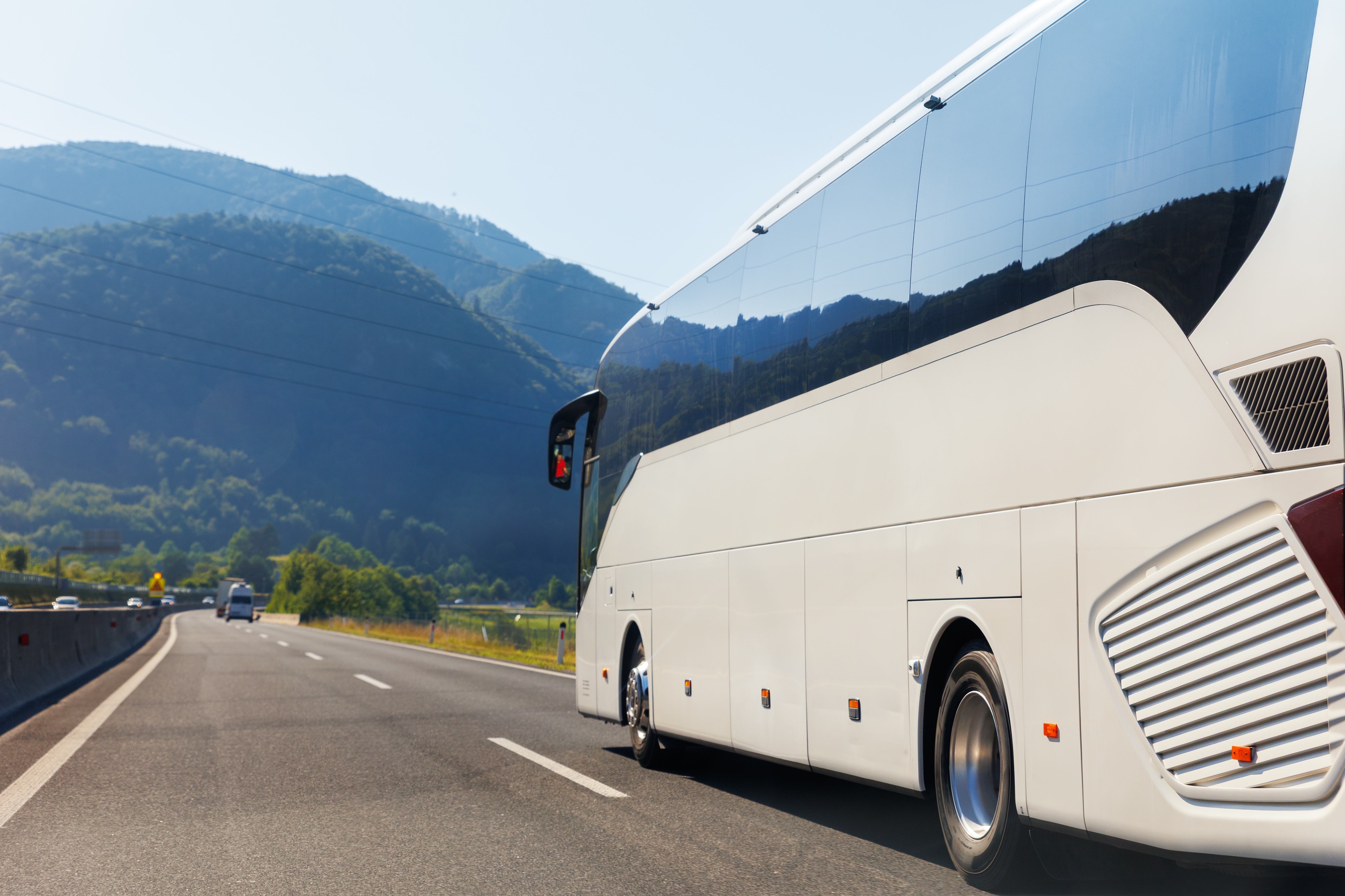 Moderne hvid turistbus kører på motorvej gennem smukt alpint bjerglandskab på en solrig sommerdag