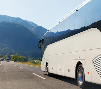 Moderne hvid turistbus kører på motorvej gennem smukt alpint bjerglandskab på en solrig sommerdag