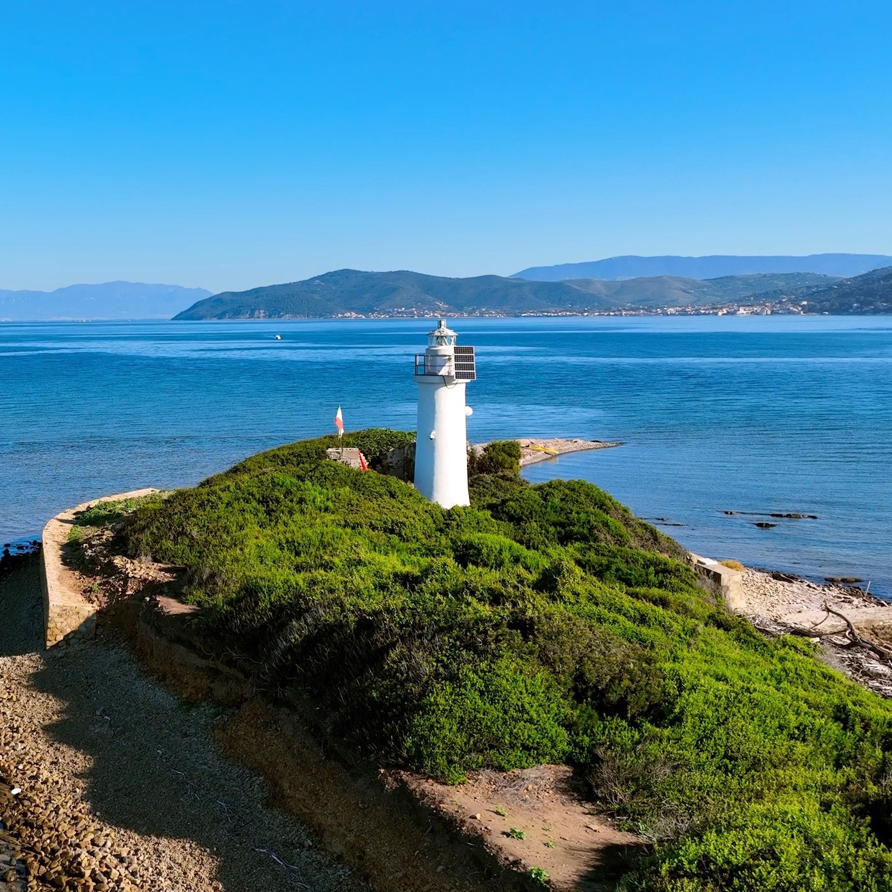 Hvidt fyrtårn på grøn ø i Middelhavet med blåt hav og bjerge