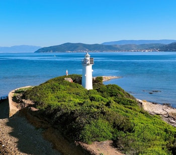 Hvidt fyrtårn på grøn ø i Middelhavet med blåt hav og bjerge