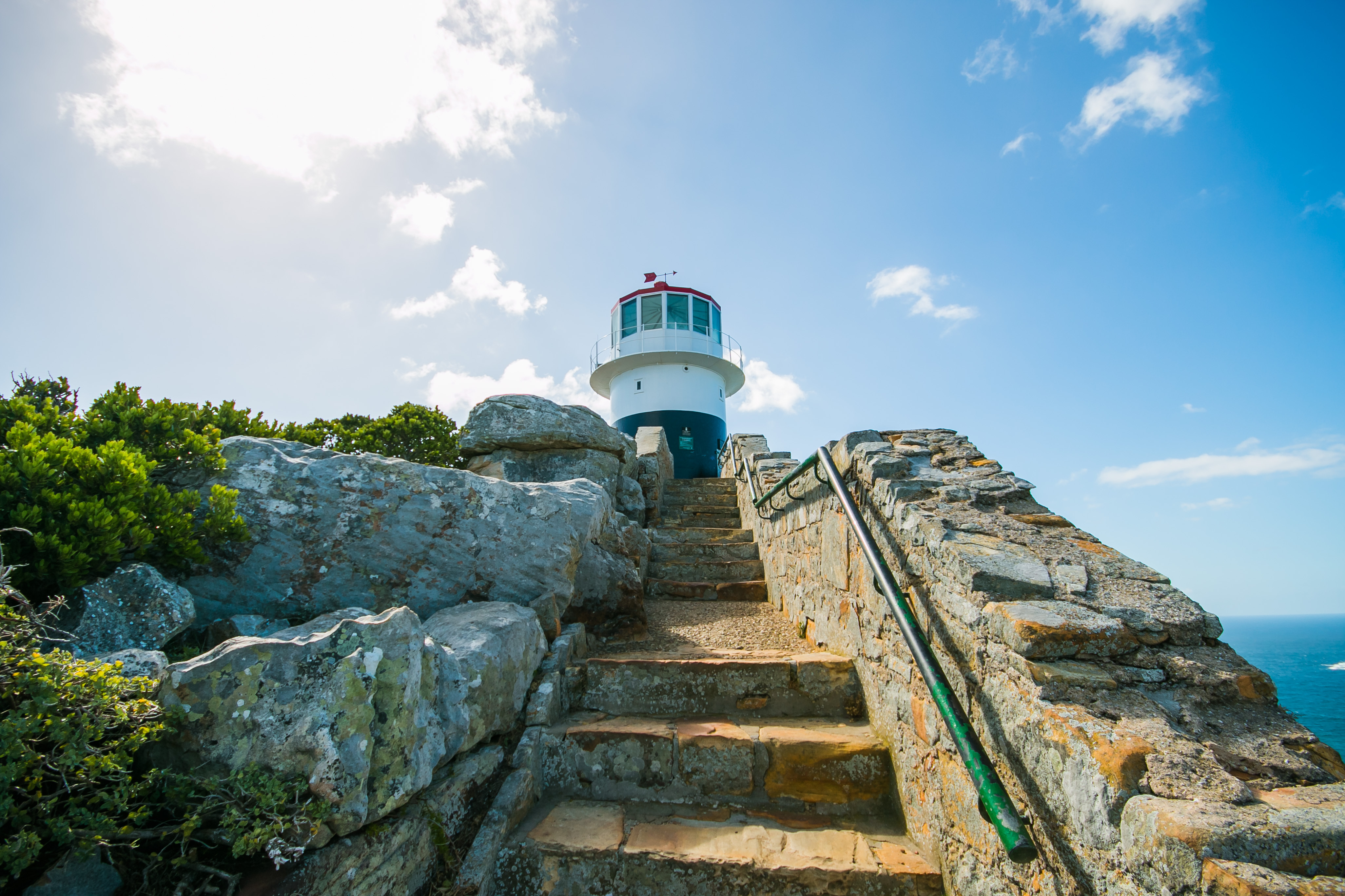 Hvidt fyrtårn på klippefyldt kyst med stentrappe og blå havudsigt