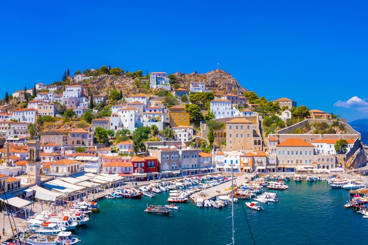 Panoramaudsigt over Hydras maleriske havn med traditionelle stenhuse, blåt vand og forankrede både - en bilfri ø i det Ægæiske Hav på vandreferie i Grækenland