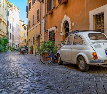 Charmerende smal gade i Trastevere-kvarteret i Rom med traditionelle italienske bygninger, udendørs caféer og frodige planter - perfekt til kulinariske oplevelser