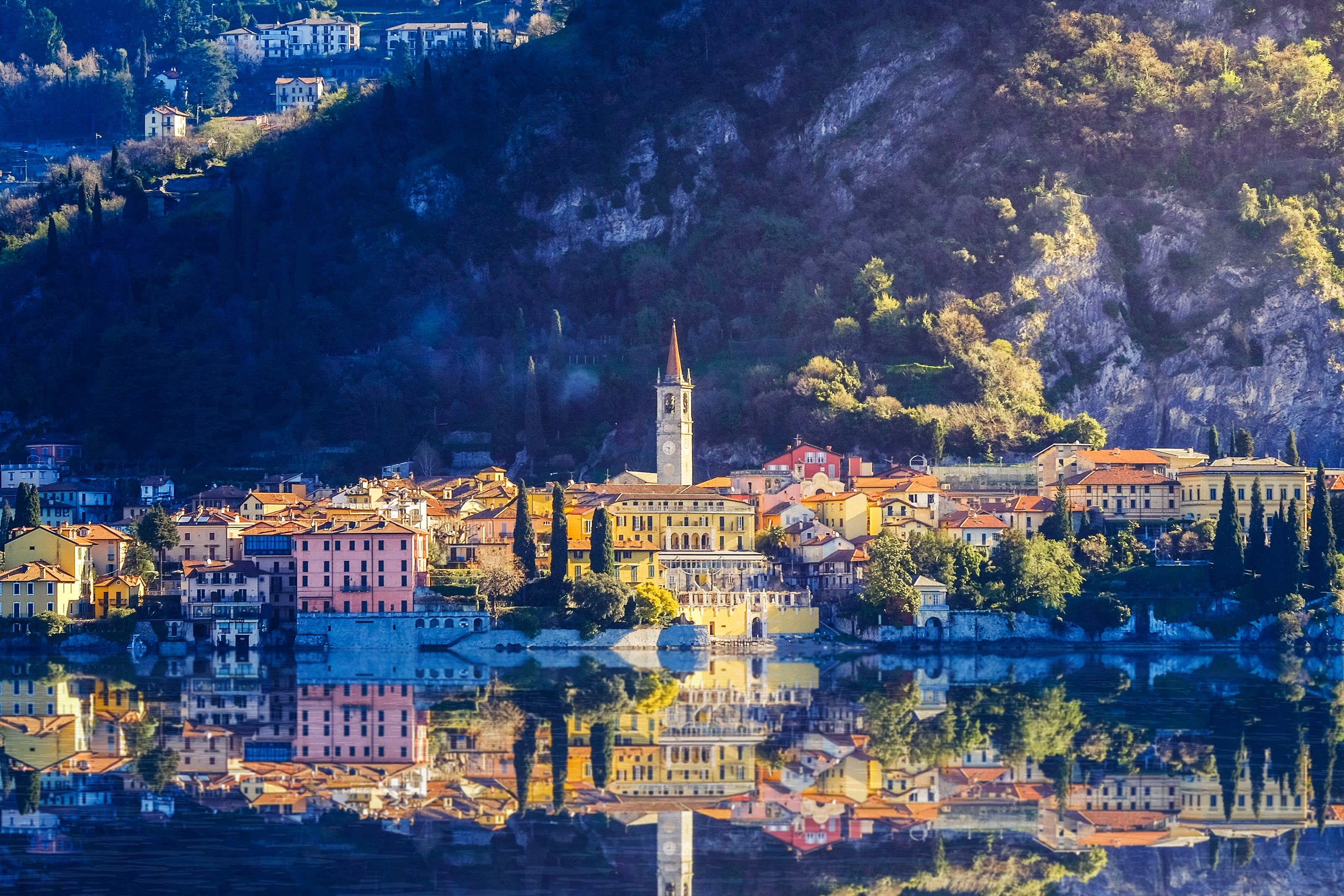 Charmerende landsby med farverige bygninger spejler sig i Como-søens blå vand med majestætiske bjerge i baggrunden, Italien