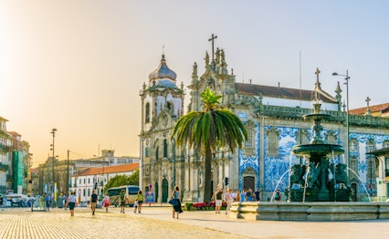 Den historiske Igreja do Carmo kirke med blå azulejo-fliser på facaden ved solnedgang i Porto, Portugal, med palmer og springvand på pladsen