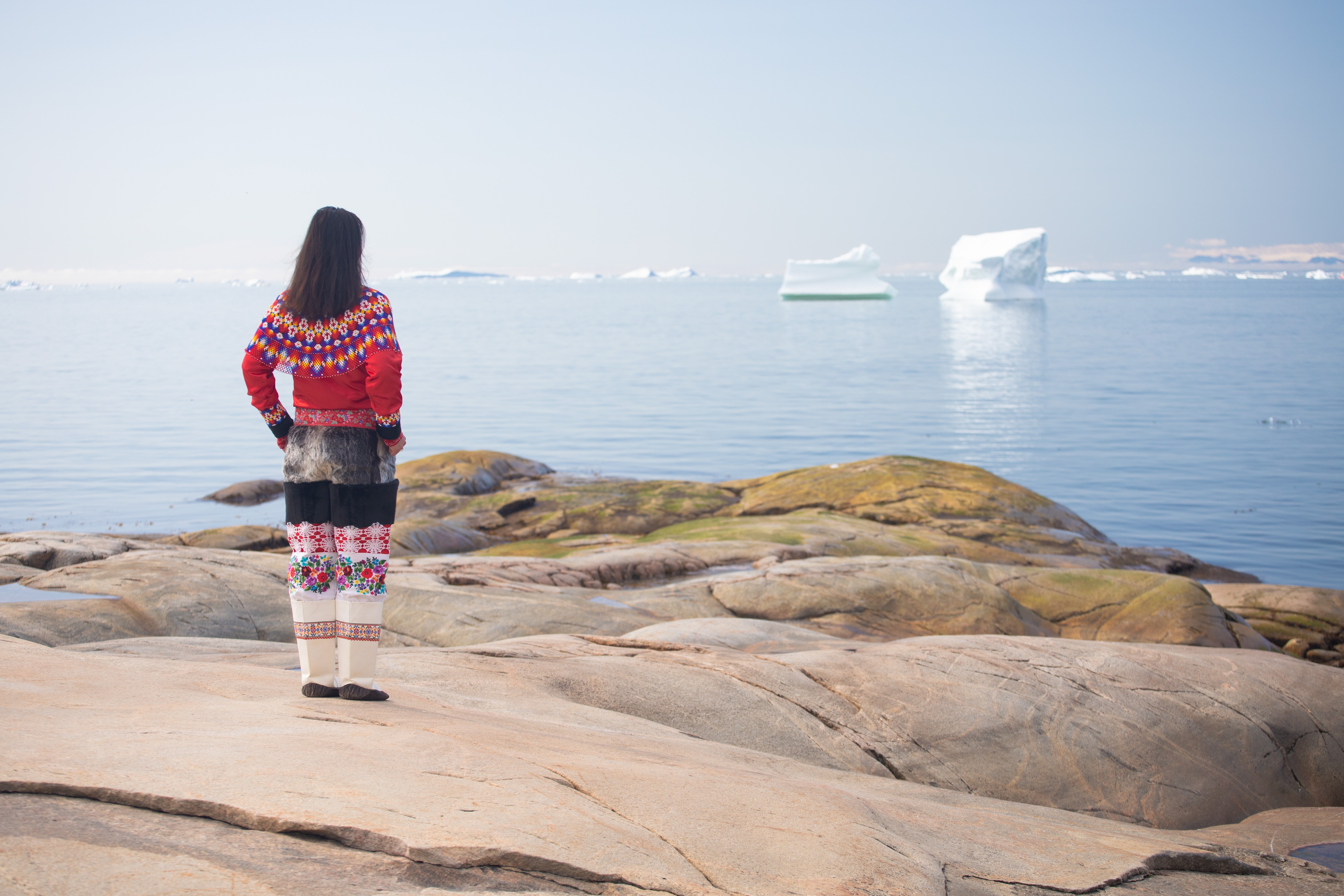 Ung inuitkvinde i farverig traditionel grønlandsk dragt står på klippekysten ved Ilulissats isfjord med isbjerge i baggrunden